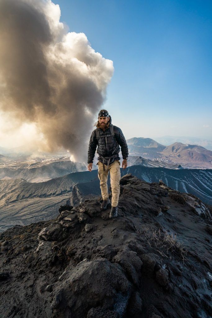 a man standing on top of a mountain next to a plume of smoke.